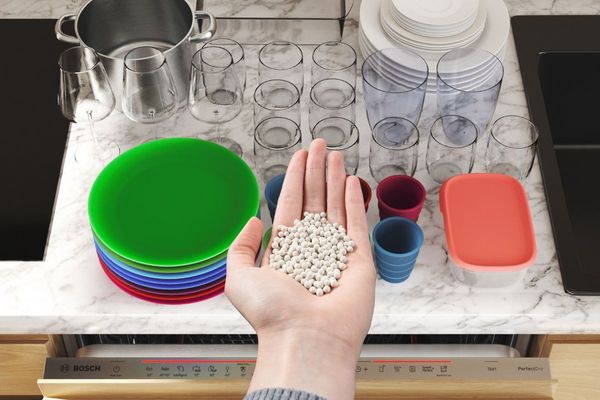 Hand met zeolietkorrels boven een open Bosch PerfectDry vaatwasser met kleurrijk serviesgoed.