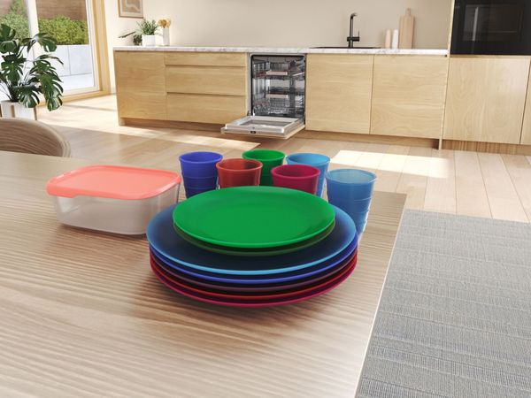 Kleurrijk kunststof serviesgoed op een eettafel met op de achtergrond een open inbouw vaatwasser.          
