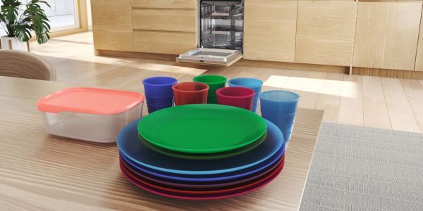 Kleurrijk kunststof serviesgoed op een eettafel met op de achtergrond een open inbouw vaatwasser.         