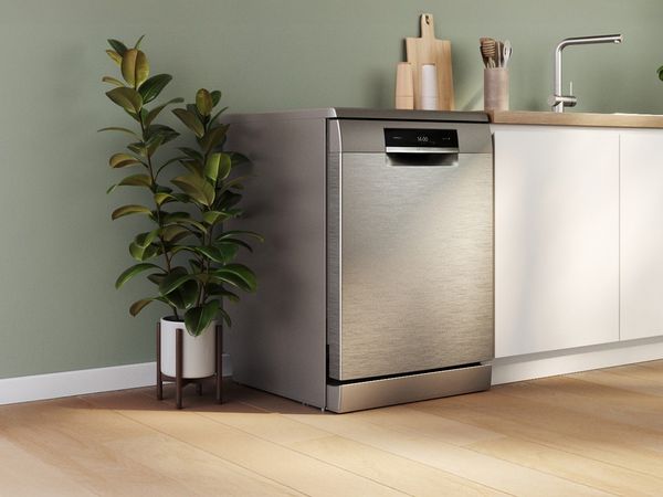 Lavastoviglie a libera installazione in acciaio inox inserita in una cucina con mobili bianchi e una pianta ornamentale