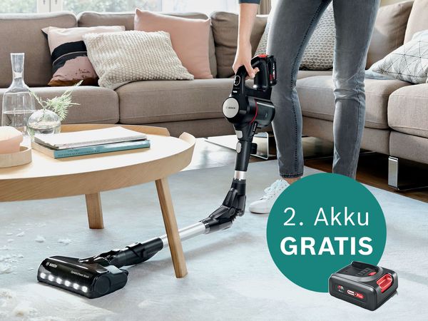 Eine Person verwendet einen kabellosen Staubsauger von Bosch auf einem mit Teppich ausgelegten Wohnzimmerboden neben einem Couchtisch und wirbt f&uuml;r das Angebot eines gratis Akku.