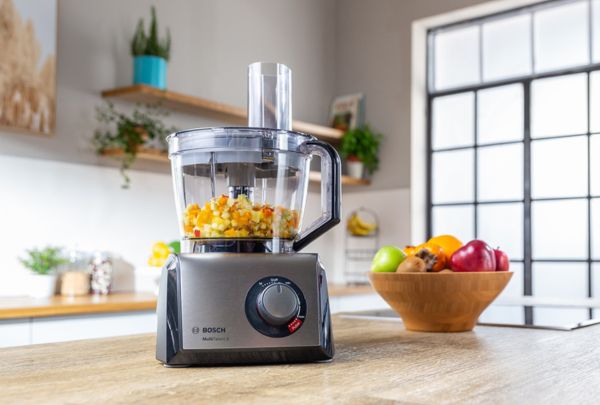 מעבד מזון של בוש עם תפוחים בתוכו מונח על שולחן לצד קערת פירות