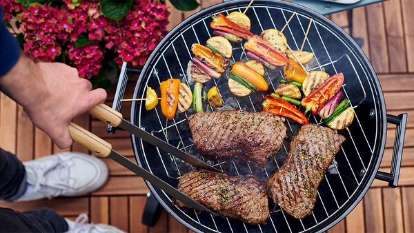 Offener Kugelgrill, auf dessen Grillrost marinierte Gem&uuml;sespie&szlig;e und marinierte Fleisch-Steaks gegrillt werden.