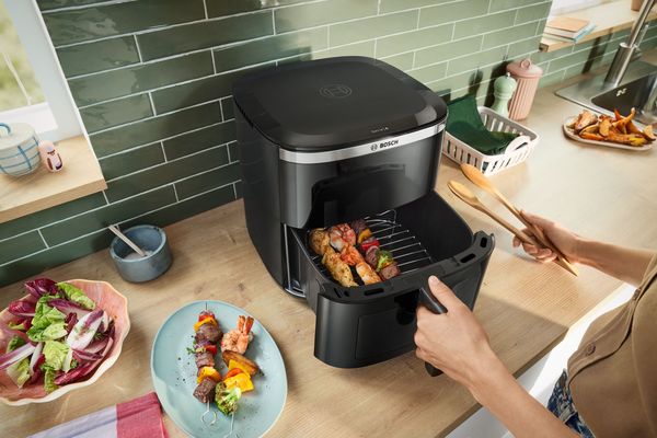 Persona che estrae il cestello di una friggitrice ad aria con spiedini di carne e verdure all&rsquo;interno