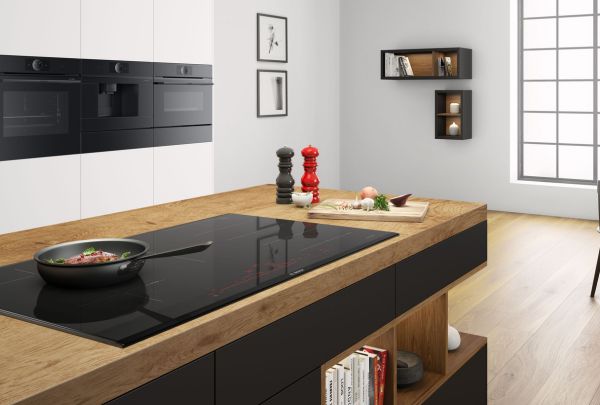 מטבח מודרני עם אי עם דלתות שחורות ושיש מעץ שעליו כיריים ועליהם מחבת. ברקע תנורי בוש