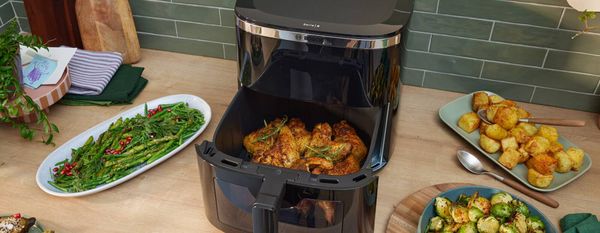 Friggitrice ad aria Bosch con cestello aperto e pollo arrosto all&rsquo;interno, circondata da contorni di patate, cavoletti e asparagi