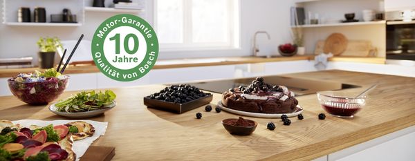 Moderne K&uuml;che mit Kochfeld, Kuchen und frischen Zutaten auf Holzarbeitsplatte, Bosch 10 Jahre Motor-Garantie Symbol.
