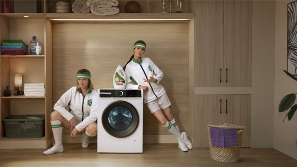 2 personnes dans une buanderie accoud&eacute;es &agrave; un lave-linge, un bidon de lessive Ariel pos&eacute; sur le lave-linge