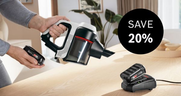 A person holds a Bosch cordless handheld vacuum cleaner and a spare battery, while another battery charges, with a "SAVE 20%" overlay.