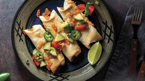 Three baked taquitos on a decorative plate topped with cucumber‑tomato salsa, sliced jalape&ntilde;o, melted cheese and a lime wedge; fork and napkin at right.