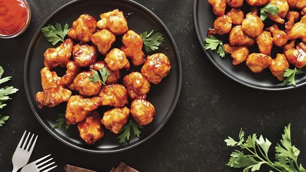 Two black plates filled with glazed cauliflower bites garnished with parsley, with a small bowl of sauce and forks on a dark surface.