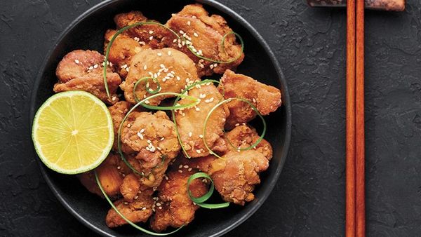 A black bowl filled with crispy fried chicken pieces, garnished with sesame seeds, green onion curls, and a lime wedge, with chopsticks on the side.