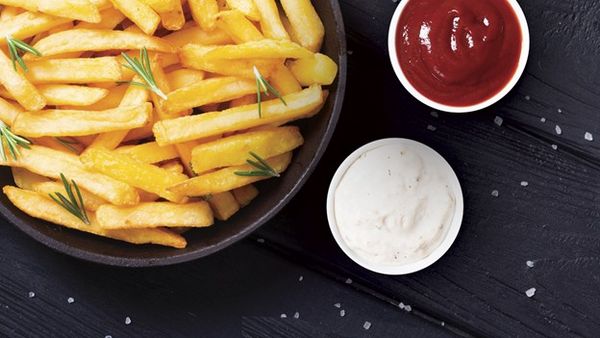 Golden French fries garnished with rosemary, served with ketchup and a creamy dip on a dark board. 
