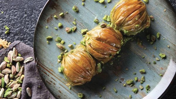 Three syrup‑glazed round pistachio pastries on a blue plate, sprinkled with crushed pistachios and loose pistachios on a cloth at left.