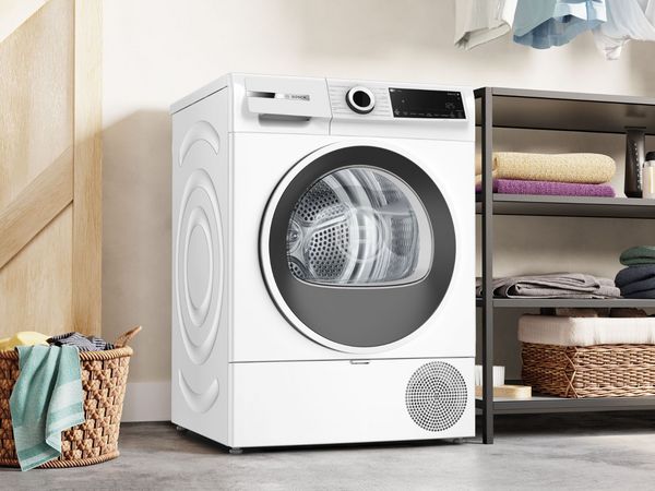 Witte Bosch warmtepompdroger in een wasruimte, met rechts een metalen rek met gevouwen handdoeken en links een rieten wasmand met een handdoek.          