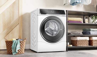 Een moderne witte wasmachine staat in een lichte wasruimte, met kledingstukken die in de buurt hangen en een plant in de hoek 