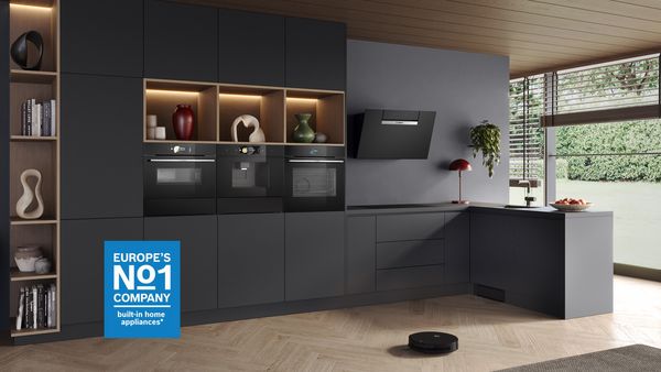 Cucina moderna con mobili scuri, forni da incasso, ripiani aperti con decorazioni, cappa nera angolata e aspirapolvere robotizzato su pavimento in legno.