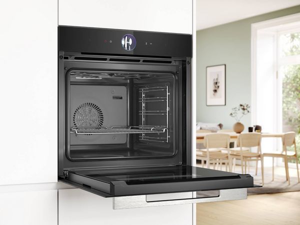 A modern black Bosch oven with its door partially open, revealing a clean interior with racks, integrated into white kitchen cabinetry, with a dining area visible in the background.