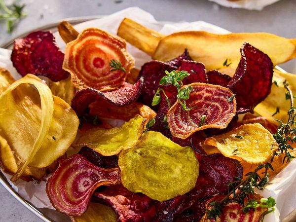 A close-up of a platter of colorful homemade vegetable chips, including red beet, golden parsnip, and yellow beet, garnished with fresh herbs.