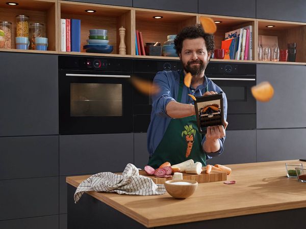 A smiling man in a green apron uses a mandoline to slice vegetables, with slices flying in the air, in a modern kitchen with integrated Bosch ovens.