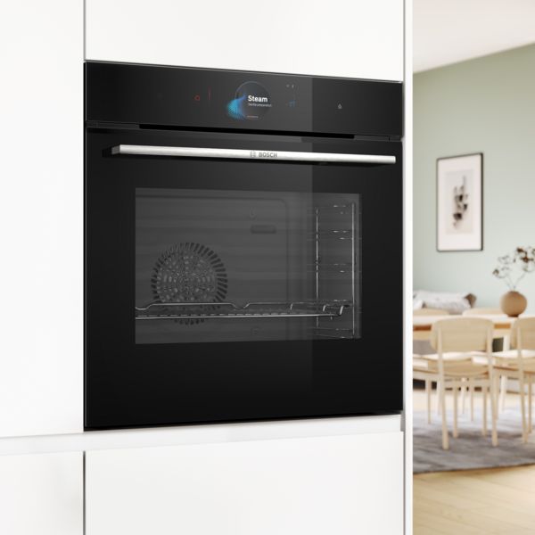 Un four encastrable noir de Bosch, int&eacute;gr&eacute; dans des meubles de cuisine blancs. L'&eacute;cran num&eacute;rique affiche la fonction de cuisson vapeur et un coin repas est visible en arri&egrave;re-plan flou.