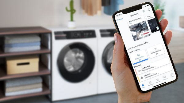 Mano che tiene uno smartphone con app Bosch per il controllo della lavatrice, con elettrodomestici sullo sfondo