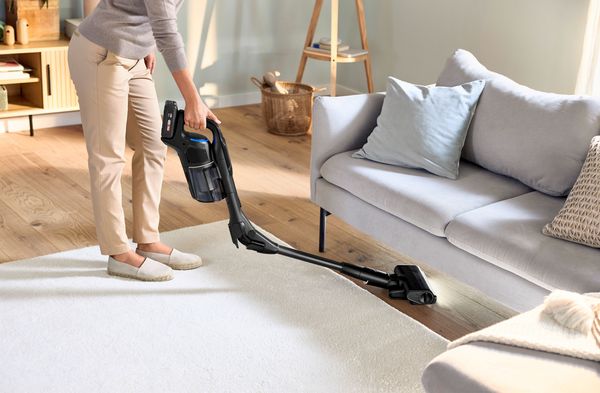 Donna che utilizza una scopa elettrica Bosch Unlimited per pulire sotto il divano in soggiorno