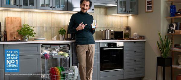 Une main tient un smartphone au premier plan, une cuisine chaleureuse se trouve en arri&egrave;re-plan.