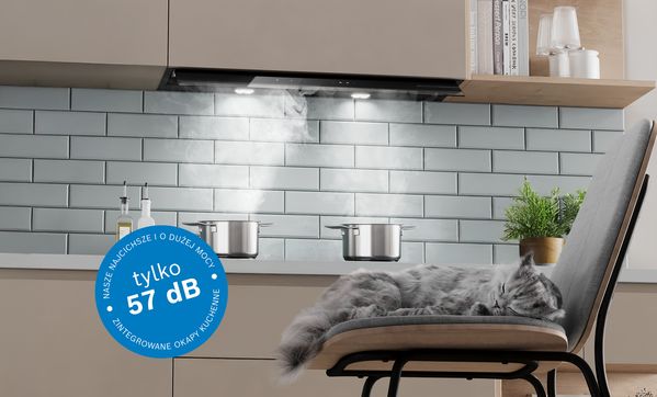 Kot śpiący przed płytą grzewczą z parującymi garnkami i zintegrowanym okapem kuchennym powyżej. Grafika odznaki poziomu hałasu 57 dB na obrazie.