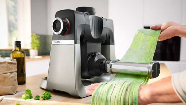 H&auml;nde f&uuml;ttern frischen gr&uuml;nen Nudelteig in eine Bosch Nudelmaschine auf der K&uuml;chenzeile, Zutaten liegen daneben.