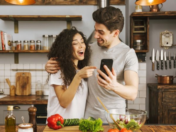 Paar in K&uuml;che zeigt gemeinsam Smartphone beim Kochen; geschnittenes Gem&uuml;se auf der Arbeitsfl&auml;che.
