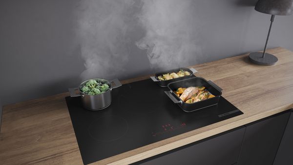 Hotte de cuisine noire encastr&eacute;e entre deux placards en bois clair, au-dessus d&rsquo;une cr&eacute;dence en carreaux rectangulaires bleu-vert ; &agrave; droite, &eacute;tag&egrave;re ouverte avec quelques livres, verres et une plante.   