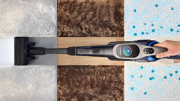 Top view of a hand holding a cordless vacuum cleaner that cleans different floor types divided into concrete, wood, dirt, and carpet sections.