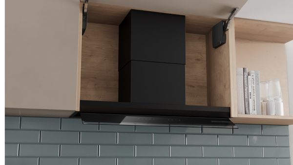 Hotte de cuisine noire encastr&eacute;e entre deux placards en bois clair, au-dessus d&rsquo;une cr&eacute;dence en carreaux bleu-vert, avec &eacute;tag&egrave;re contenant livres, verres et plante.   