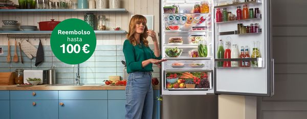 Mujer en una cocina junto a un frigor&iacute;fico abierto y organizado, lleno de alimentos frescos, con la promoci&oacute;n "Reembolso hasta 100 &euro;".