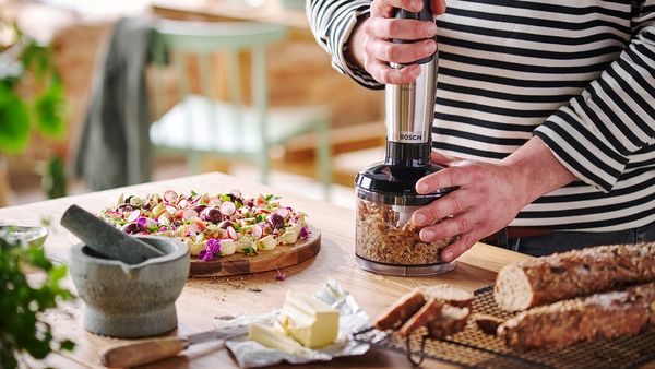 Ein Mann steht am K&uuml;chentresen und zerkleinert f&uuml;r ein Butter Board Waln&uuml;sse mit einem ErgoMaster Serie 6 Stabmixer von Bosch inklusive Universalzerkleinerer.