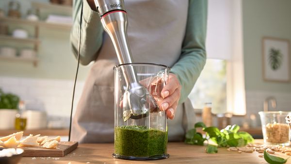 Eine Person bereitet mit einem Stabmixer gr&uuml;nes Pesto in einem Mixbecher zu.