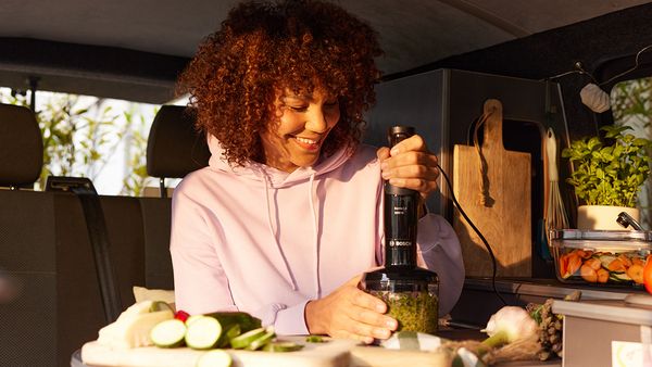 Eine dunkelhaarige Frau mit Locken und rosa Hoodie sitzt in einem Campingbus und zerkleinert Gem&uuml;se im Universal-Zerkleinerer des Bosch ErgoMaster Serie 4.