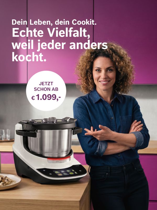 Eine l&auml;chelnde Frau mit lockigen Haaren pr&auml;sentiert in einer modernen, lila K&uuml;che die Cookit K&uuml;chenmaschine.