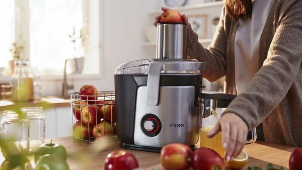 Eine Frau gibt mit einer Hand einen Apfel in den Zentrifugalentsafter VitaJuice von Bosch. Gleichzeitig greift sie mit der anderen Hand nach einem weiteren Apfel, der vor ihr liegt.