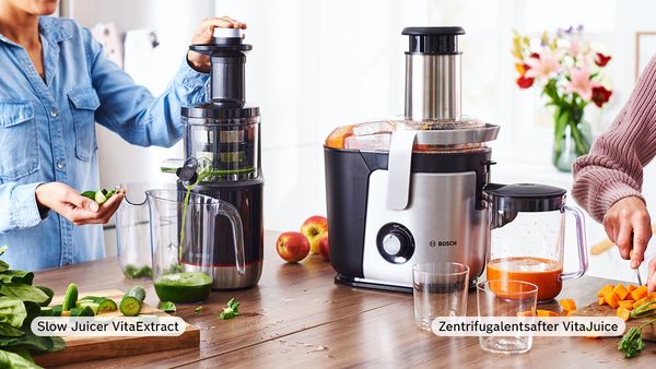 Links gibt eine Person im Jeanshemd kleingeschnittene Gurken in den Bosch VitaExtract Slow Juicer. Daneben steht ein VitaJuice Zentrifugalentsafter vor Bosch. Rechts ist die Hand einer weiteren Person zu sehen, die auf einem Holzbrett Karotten kleinschneidet.
