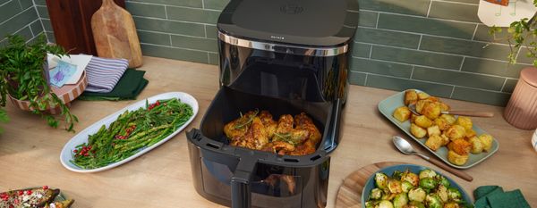 Friggitrice ad aria aperta su un piano cucina con pollo nel cestello, circondata da contorni di verdure e patate arrosto