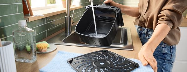 Persona che risciacqua un cestello nero per friggitrice ad aria sotto l'acqua corrente in un moderno lavello da cucina con paraschizzi piastrellato di verde. 