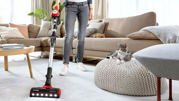 Eine Frau saugt mit einem Bosch Unlimited 7 Staubsauger den Wohnzimmerteppich, w&auml;hrend eine Katze auf einem Sitzsack liegend zusieht.