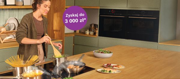 Nowoczesna kuchnia z czarnymi urządzeniami, drewnianą wyspą i jasnym wystrojem. Baner promuje cashback do 1800 zł.