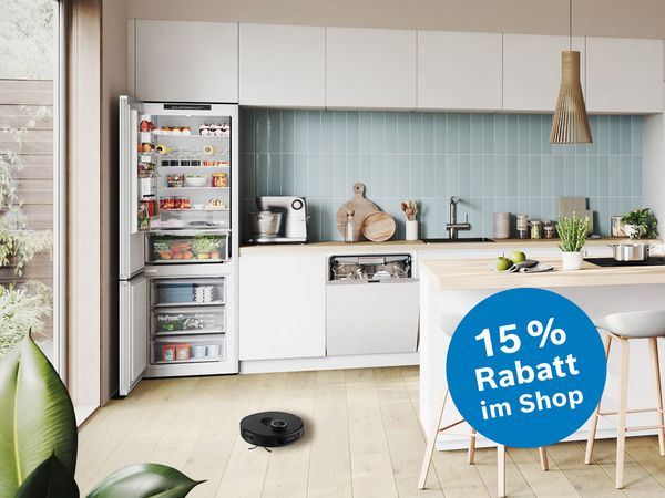 Moderne K&uuml;che mit offenem K&uuml;hlschrank, Geschirrsp&uuml;ler, Kaffeemaschine, Esstisch aus Holz und einem Staubsaugerroboter auf hellem Holzboden.