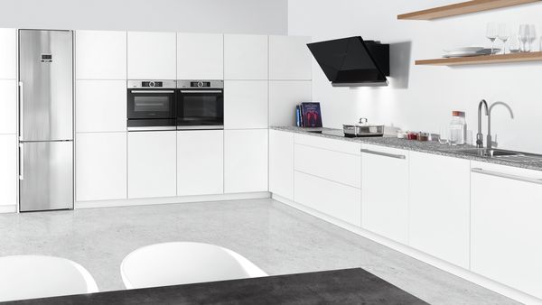 Large, L-shaped open kitchen with modern white cabinets. Built-in Bosch twin ovens. Fridge freezer in stainless steel. Wall-mounted cooker hood above hob at right. Open shelving with minimalist tableware. Grey flooring in concrete look. Table corner echoes the contours of kitchen. 