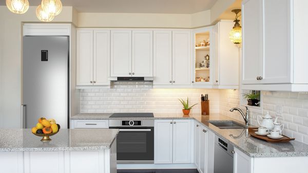 Separate medium-sized kitchen with island. White farmhouse cabinetry, marble countertops. Bosch fridge and dishwasher with stainless steel fronts. Black Bosch oven. Bowl of fruit on island. Tea set on counter at right. Small plants and a knife block in background. 
