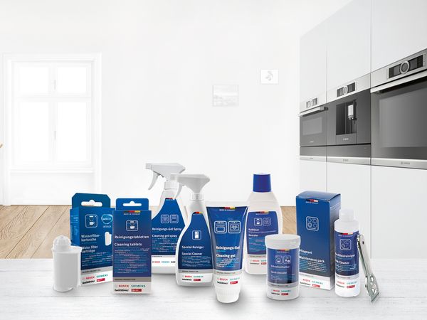 Reinigungs- und Pflegeprodukte f&uuml;r Haushaltsger&auml;te (Bosch/Siemens) auf Holztisch vor moderner K&uuml;che.