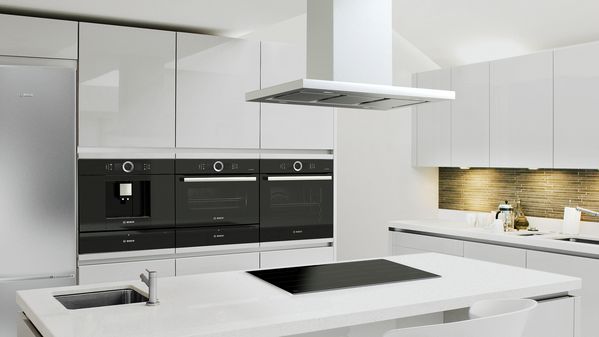 Modern ergonomic kitchen with twin ovens and a fully automated espresso maker at eye level. Sleek white cabinetry, centre island with flush-fitting hob and ceiling-mounted cooker hood. 